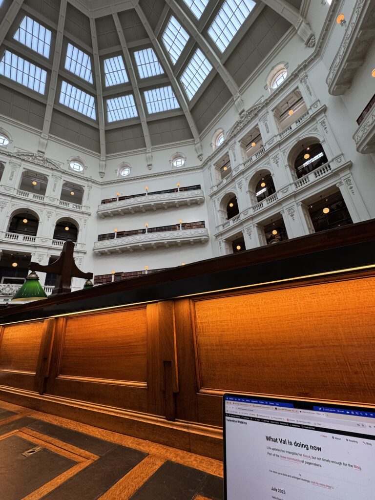 Wide shot of the State Library of Victoria, beneath the grand dome. Despite the grandeur, the wood-panelled reading desks are cosily-lit.